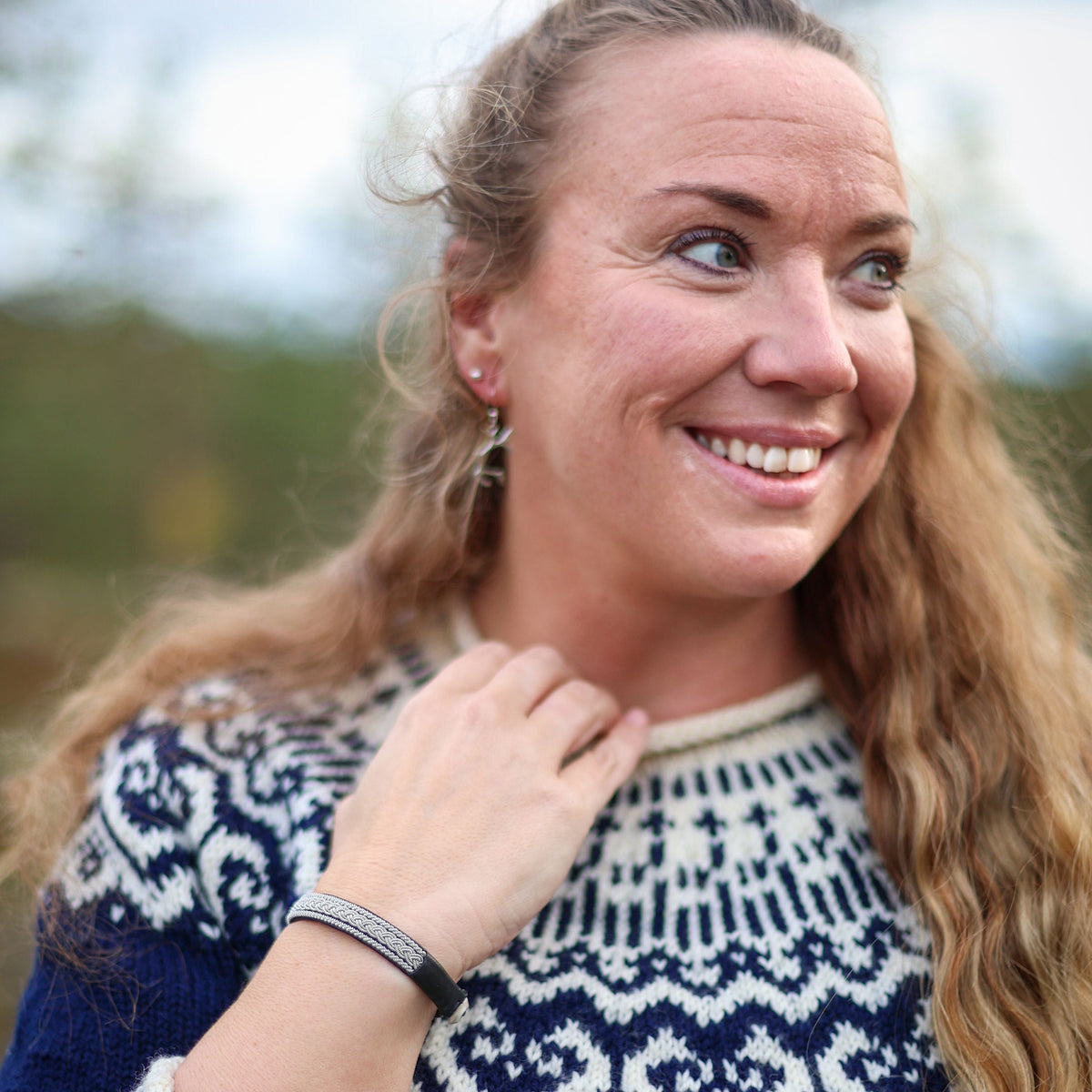 A smiling woman with long brown hair wearing a navy blue and white knit sweater, photographed outdoors with a Frost pewter and leather bracelet