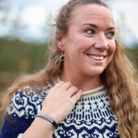 A smiling woman with long brown hair wearing a navy blue and white knit sweater, photographed outdoors with a Frost pewter and leather bracelet