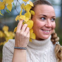 A woman with blonde hair in a braid wearing a cream sweater, standing outdoors in a forest with yellow autumn leaves in the foreground wearing a leather bracelet.
