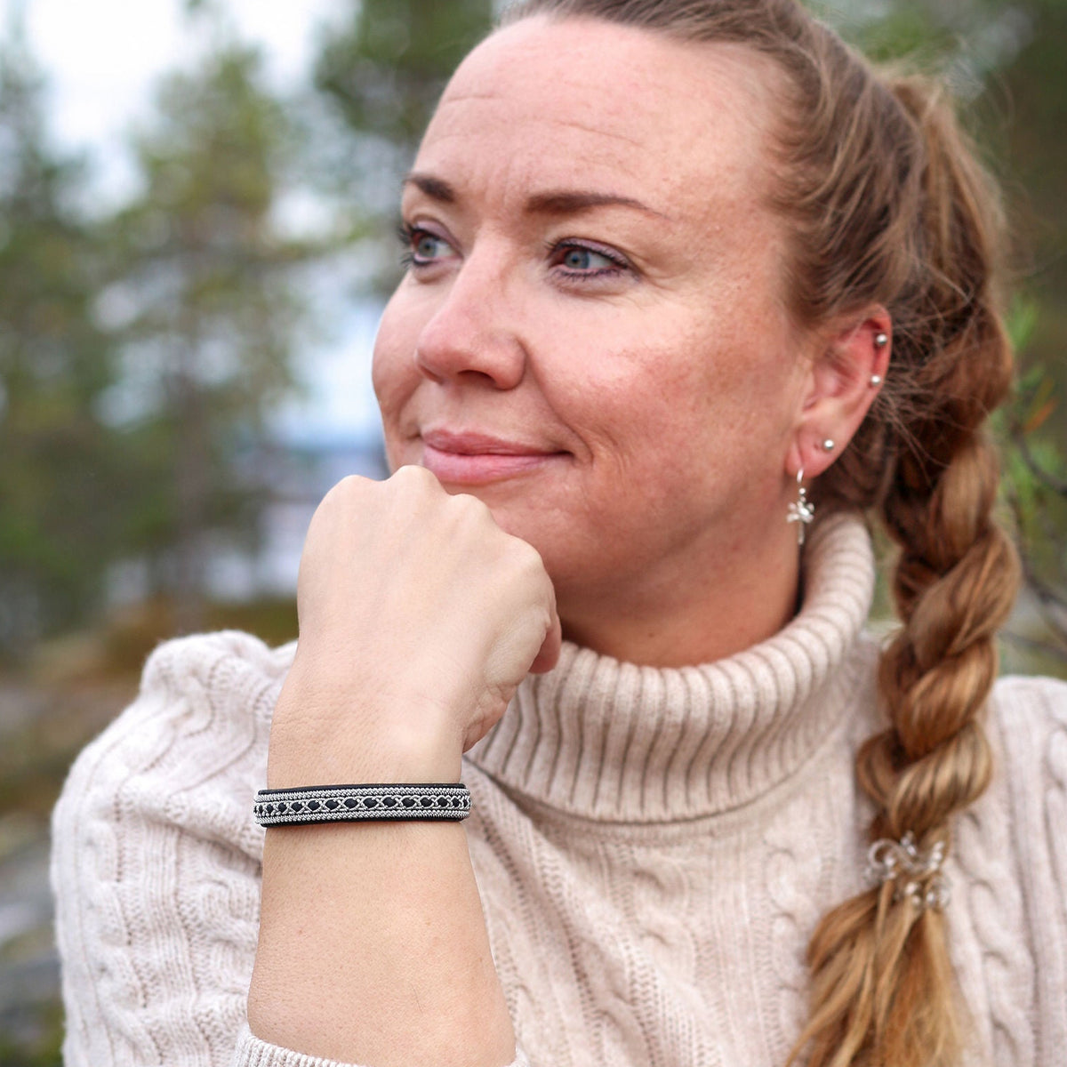 A woman with blonde hair in a braid wearing a cream cable-knit turtleneck sweater, wearing a black sami bracelet in an outdoor forest setting.