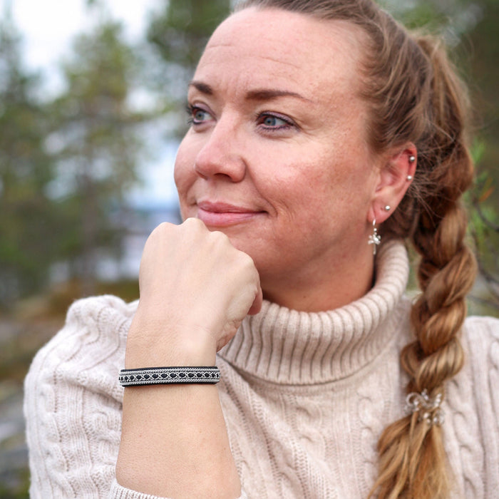 A woman with blonde hair in a braid wearing a cream cable-knit turtleneck sweater, wearing a black sami bracelet in an outdoor forest setting.